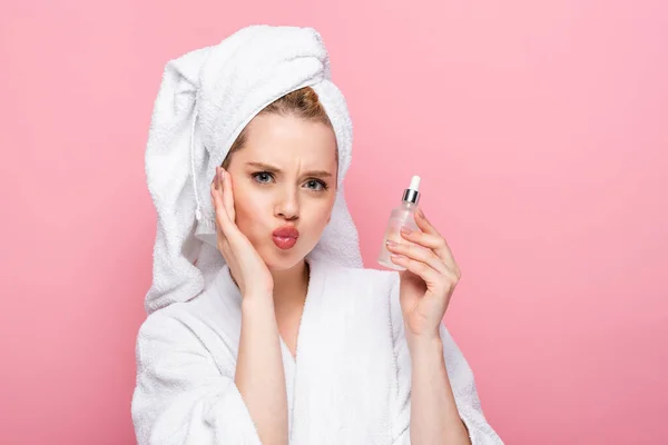 Young woman in bathrobe with towel on head holding serum and pouting lips isolated on pink — Stock Photo