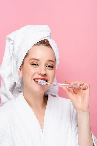 Smiling young woman in bathrobe with towel on head brushing teeth isolated on pink — Stock Photo