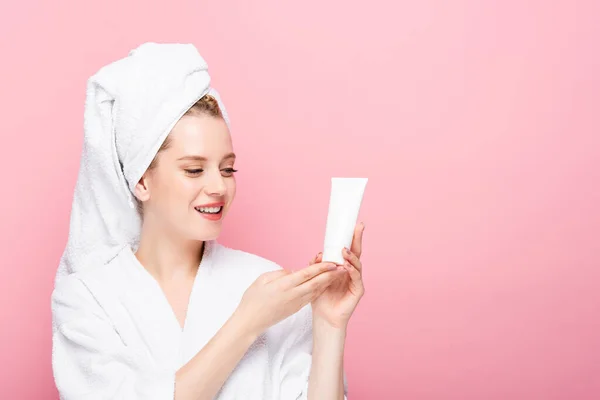 Happy young woman in bathrobe with towel on head holding tube with hand cream isolated on pink — Stock Photo