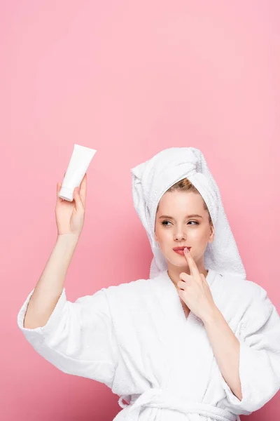 Chère jeune femme en peignoir avec serviette sur la tête tenant tube avec crème pour les mains isolée sur rose — Photo de stock