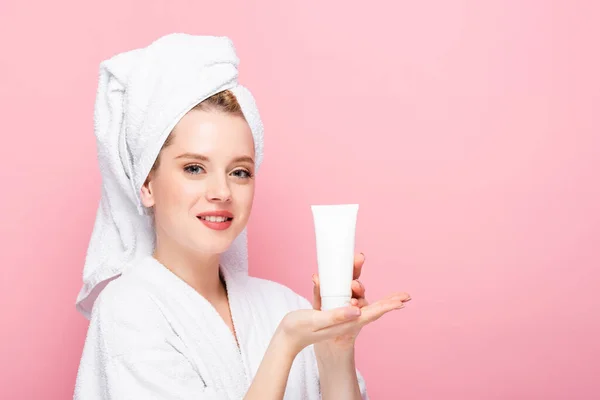 Young woman in bathrobe with towel on head holding tube with hand cream isolated on pink — Stock Photo