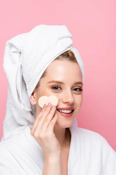 Happy young woman in bathrobe with towel on head and facial heart shaped sponge isolated on pink — Stock Photo