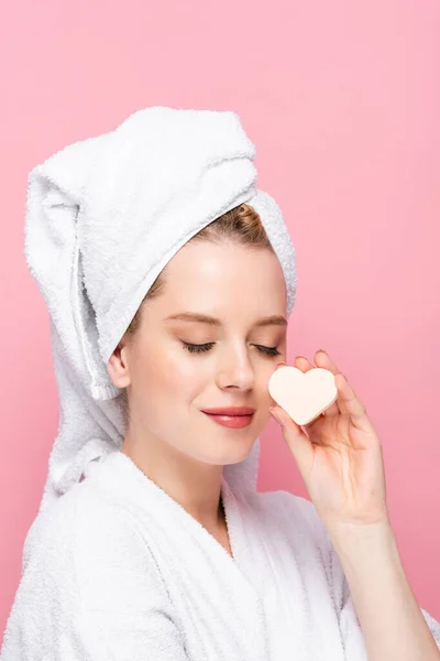 Mujer joven en albornoz con los ojos cerrados, toalla en la cabeza y esponja en forma de corazón facial aislada en rosa - foto de stock