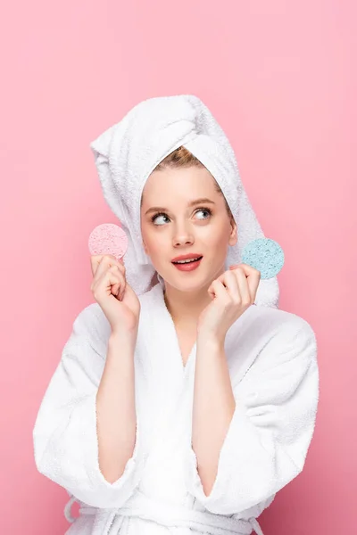 Jovem sonhadora em roupão de banho com toalha na cabeça segurando esponjas faciais isolados em rosa — Fotografia de Stock