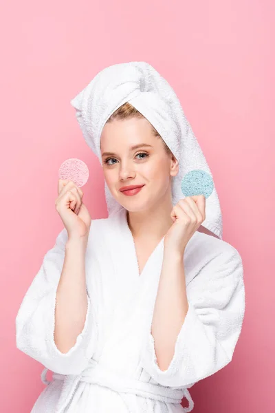 Jeune femme en peignoir avec serviette sur la tête tenant des éponges faciales isolées sur rose — Photo de stock