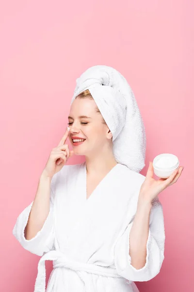 Щаслива молода жінка в рушнику на голові тримає косметичний крем і наносить його на ніс ізольовано на рожевий — стокове фото