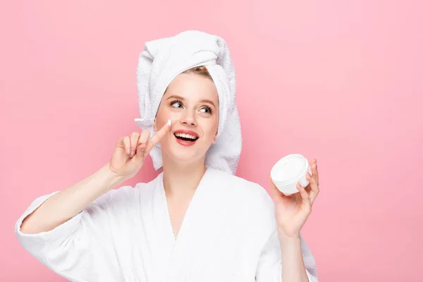 Glücklich junge Frau im Bademantel Handtuch auf dem Kopf hält kosmetische Creme und zeigt den Finger isoliert auf rosa — Stockfoto