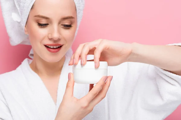 Jeune femme en peignoir serviette sur la tête ouverture crème cosmétique isolé sur rose — Photo de stock