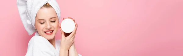 Happy young woman in bathrobe with closed eyes, towel on head holding cosmetic cream isolated on pink, banner — Stock Photo