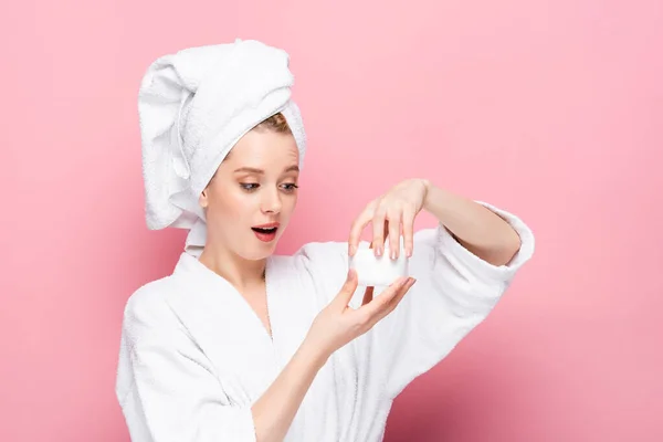 Shocked young woman in bathrobe with towel on head opening cosmetic cream isolated on pink — Stock Photo