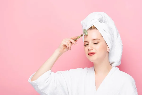 Jeune femme en peignoir avec serviette sur la tête en utilisant un rouleau de jade isolé sur rose — Photo de stock