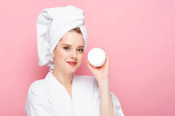 Jovem mulher em roupão de banho com toalha na cabeça segurando creme cosmético isolado em rosa — Fotografia de Stock