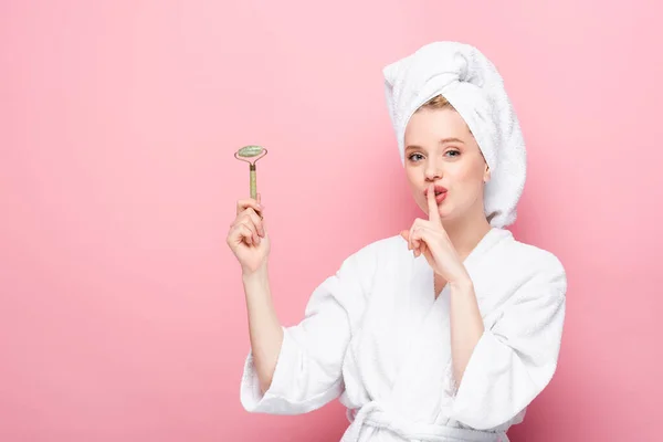 Young woman in bathrobe with towel on head holding jade roller and showing shh isolated on pink — Stock Photo