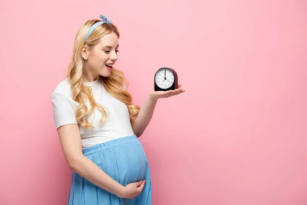 Happy blonde young pregnant woman holding watch on pink background — Stock Photo