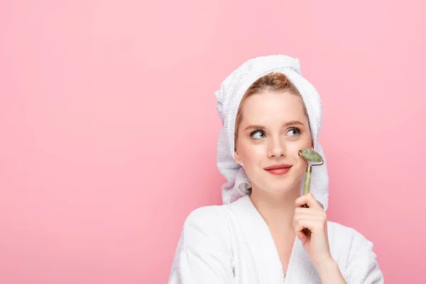 Dreamy young woman in bathrobe with towel on head using jade roller isolated on pink — Stock Photo