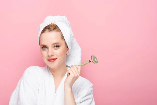 Young woman in bathrobe with towel on head and jade roller isolated on pink — Stock Photo