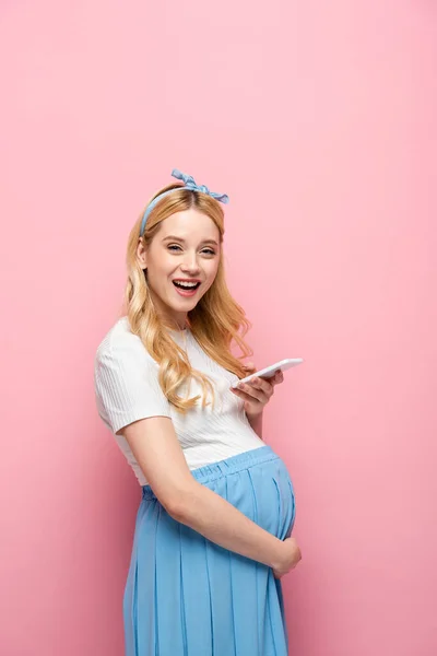 Feliz rubia joven embarazada utilizando teléfono inteligente sobre fondo rosa - foto de stock