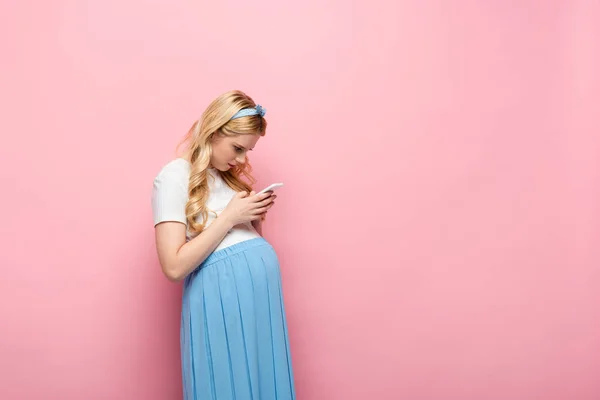 Blonde jeune femme enceinte en utilisant smartphone sur fond rose — Photo de stock