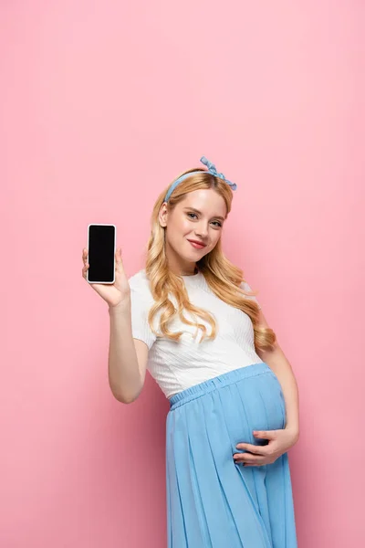Felice bionda giovane donna incinta mostrando smartphone su sfondo rosa — Foto stock