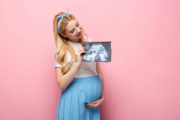 Happy blonde young pregnant woman with baby ultrasound scan on pink background — Stock Photo