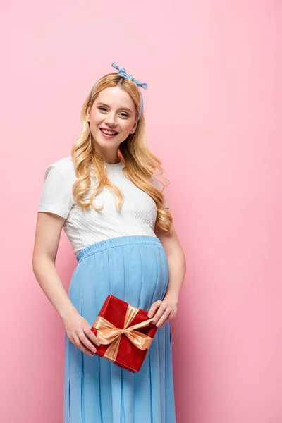 Happy blonde young pregnant woman with gift box on pink background — Stock Photo