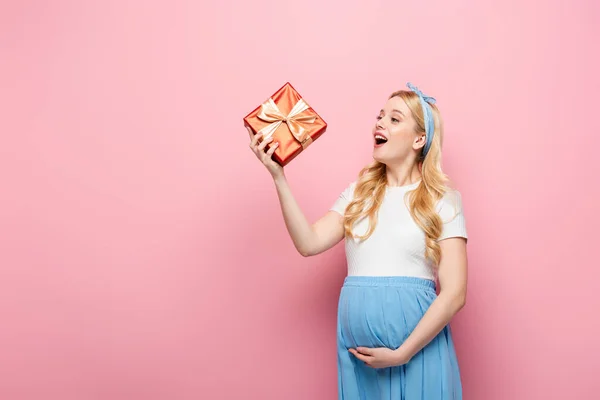Feliz loira jovem grávida com caixa de presente no fundo rosa — Fotografia de Stock