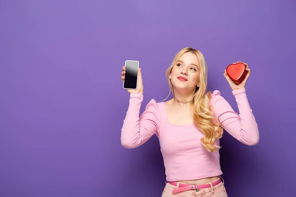 Blonde young woman with red heart shaped box and smartphone on purple background — Stock Photo