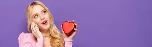 Блондинка молодая женщина с красным сердцем форме коробки говорить на смартфоне на фиолетовом фоне, баннер — стоковое фото