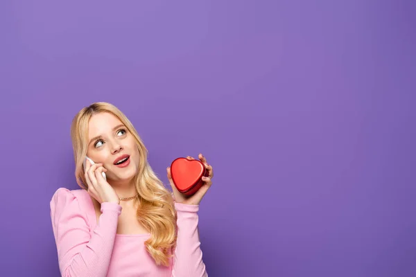 Blonde young woman with red heart shaped box talking on smartphone on purple background — Stock Photo