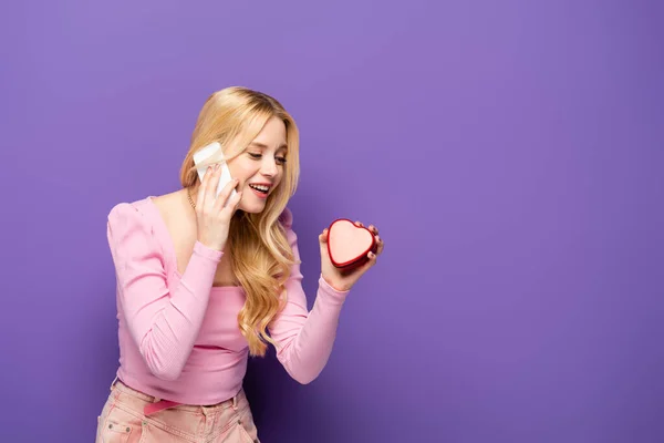 Щаслива блондинка молода жінка з червоною коробкою у формі серця розмовляє на смартфоні на фіолетовому фоні — стокове фото