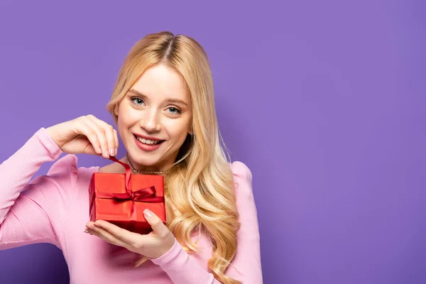Happy blonde young woman opening red gift box on purple background — Stock Photo