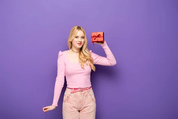 Щаслива блондинка молода жінка тримає червону подарункову коробку на фіолетовому фоні — стокове фото