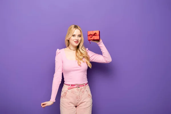 Feliz loira jovem segurando caixa de presente vermelho no fundo roxo — Fotografia de Stock