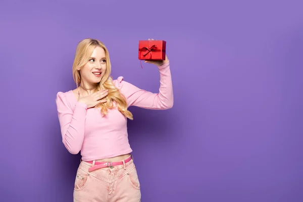 Happy blonde young woman holding red gift box on purple background — Stock Photo