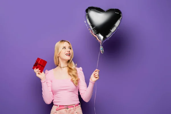 Happy blonde young woman holding heart shaped balloon and gift on purple background — Stock Photo