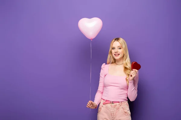 Sorridente giovane donna bionda che tiene palloncino a forma di cuore e regalo su sfondo viola — Foto stock