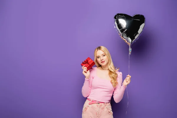 Blonde young woman holding heart shaped balloon and gift on purple background — Stock Photo