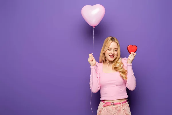 Happy blonde young woman holding red heart shaped box and balloon on purple background — Stock Photo