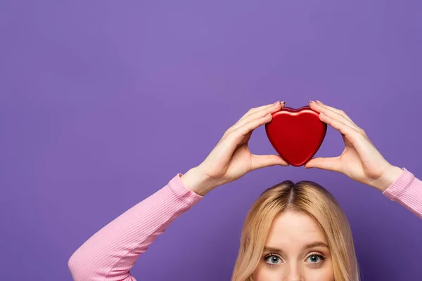 Cropped view of blonde young woman holding red heart shaped box on head on purple background — Stock Photo