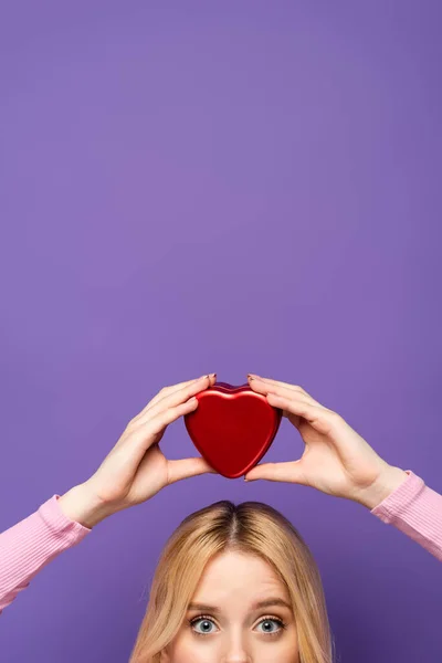 Vista recortada de la joven rubia sosteniendo caja en forma de corazón rojo en la cabeza sobre fondo púrpura - foto de stock