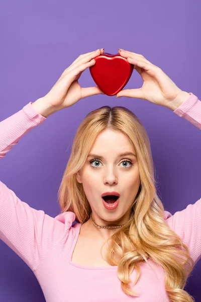 Shocked blonde young woman holding red heart shaped box above head on purple background — Stock Photo