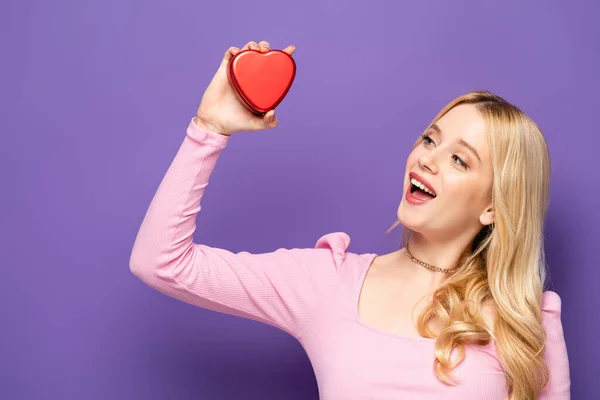 Happy blonde young woman holding red heart shaped box on purple background — Stock Photo