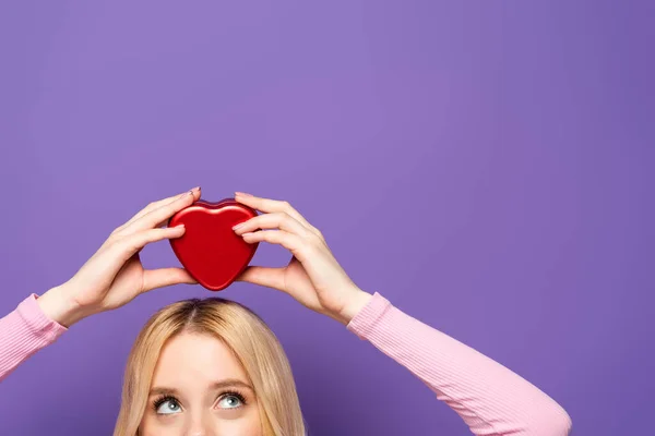 Обрезанный вид блондинка молодая женщина держит красное сердце форме коробки на голове на фиолетовом фоне — стоковое фото