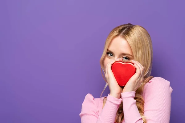 Mujer joven rubia sosteniendo caja en forma de corazón rojo cerca de la cara sobre fondo púrpura - foto de stock