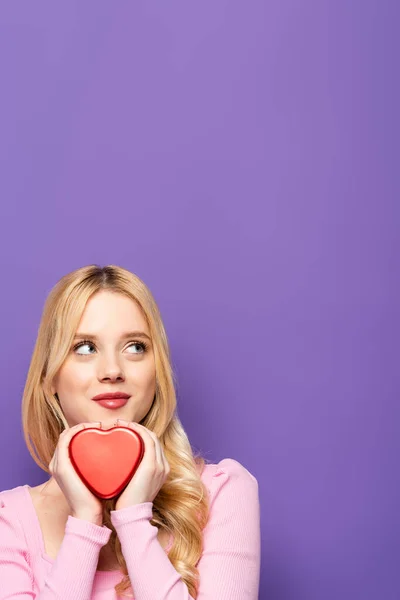 Dreamy blonde young woman holding red heart shaped box on purple background — Stock Photo