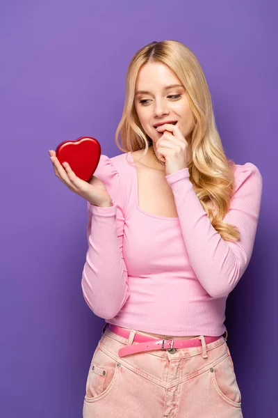 Shy blonde young woman holding red heart shaped box on purple background — Stock Photo