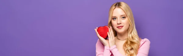 Blonde young woman holding red heart shaped box isolated on purple, banner — Stock Photo