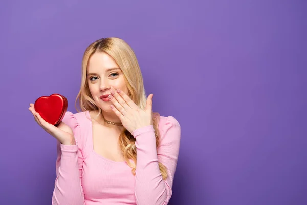 Schüchterne blonde junge Frau mit roter Herzschachtel auf lila Hintergrund — Stockfoto
