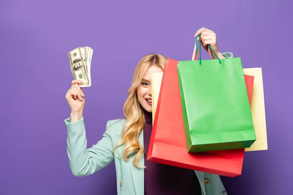 Sonriente joven rubia en blazer turquesa de moda con bolsas de compras y dinero sobre fondo púrpura - foto de stock