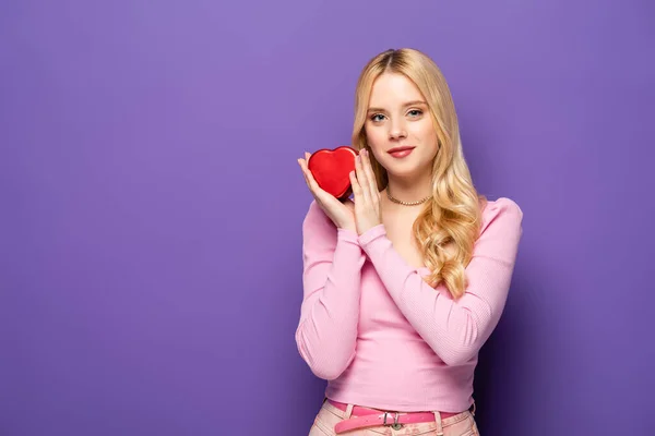 Блондинка молода жінка тримає червону коробку у формі серця на фіолетовому фоні — стокове фото